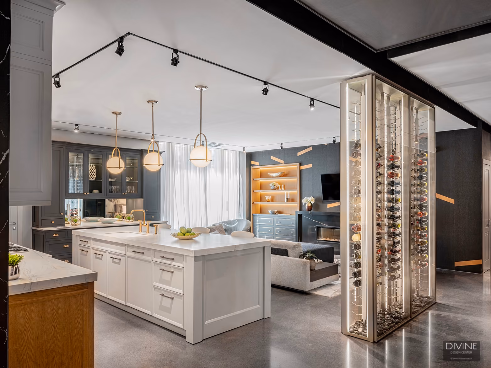Modern open-concept kitchen with a large white island featuring a bowl of green apples and three contemporary pendant lights overhead, adjacent to a cozy living area with a gray sofa, built-in wooden shelves illuminated with warm lighting, a wall-mounted TV, and a sleek glass-enclosed wine rack on a polished concrete floor.