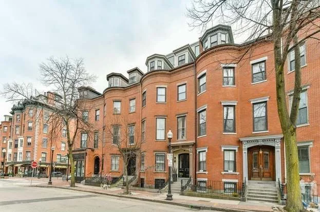 Row of classic red-brick townhouses with bay windows and dormers, lined along a quiet street with bare trees and street lamps.