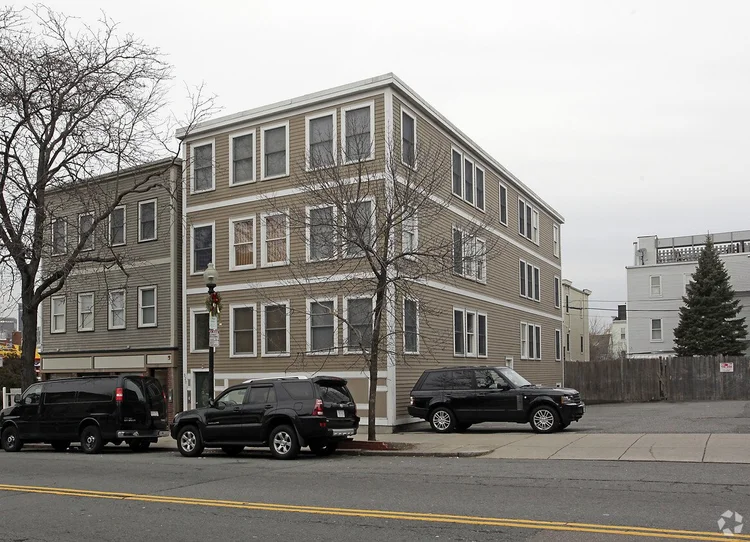 A three-story beige residential building with white trim and multiple windows on each floor, located on a corner with several parked black vehicles along the street and driveway, under a gray overcast sky.