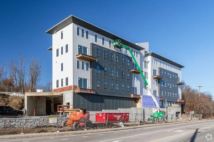 Four-story modern building under construction with white and gray siding and multiple windows, a green construction lift extended to the upper floors, red construction dumpster, and fenced perimeter along a roadside.