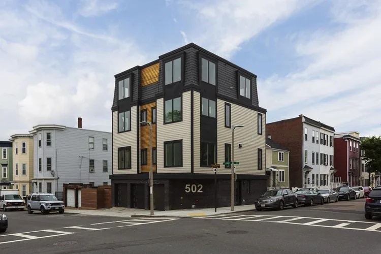 Three-story modern residential building with black and beige exterior, large windows, and the number 502 displayed on the corner, located at an urban street intersection with parked cars and neighboring older houses.