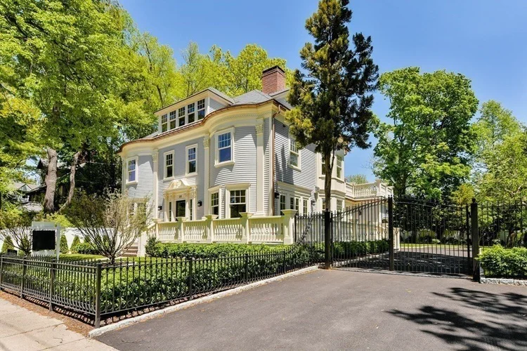 Large two-story white house with classical architectural details, surrounded by green trees and bushes, featuring wrought iron gate and fence along a paved driveway under a clear blue sky.