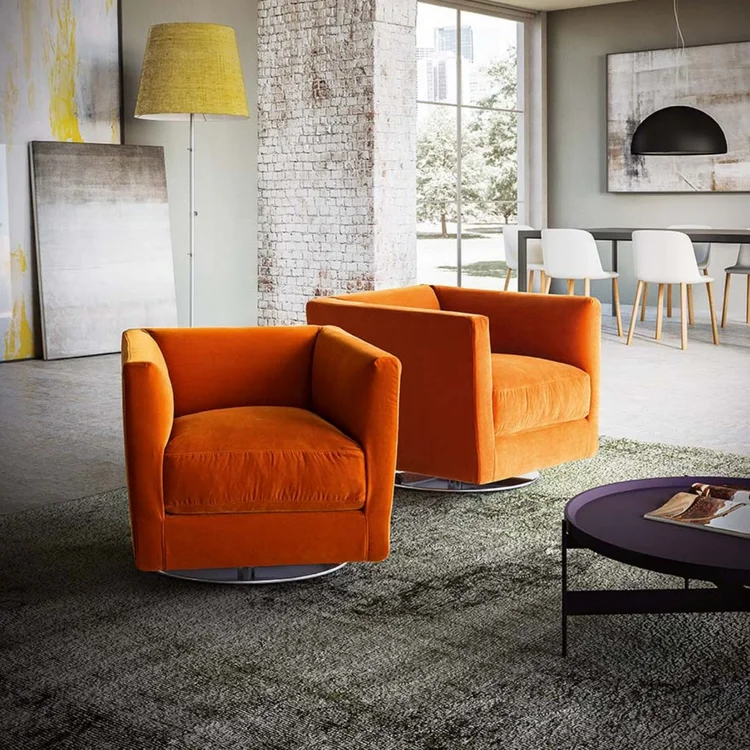 Modern living room with two vibrant orange armchairs on a green textured rug, a black round coffee table with magazines, a yellow floor lamp, and large windows showing outdoor greenery.