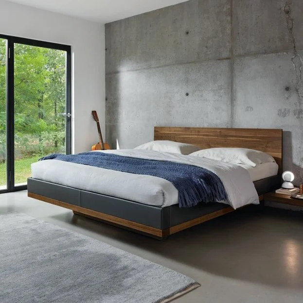 Modern bedroom with a low platform bed featuring a wooden headboard and dark upholstered frame, neatly made with white bedding and a blue throw blanket, next to a concrete wall, a small nightstand with a round lamp, and a large glass door showing green outdoor trees.