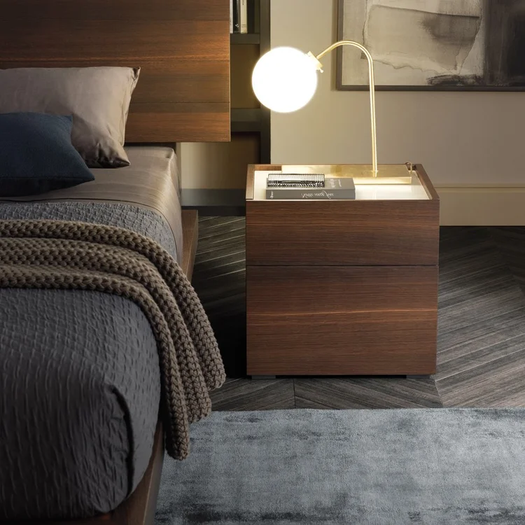 Modern bedroom corner with a wooden nightstand holding a round illuminated lamp and stacked books, next to a bed with gray bedding and a textured beige throw blanket, set on a gray carpet and herringbone wooden floor.
