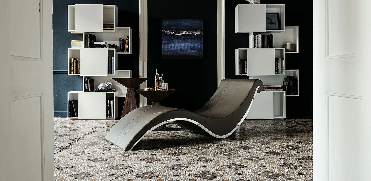 Modern curved lounge chair in dark brown leather with white base positioned on patterned tile floor, flanked by white modular bookshelves and dark wooden side tables with glassware, against a black wall with a square abstract artwork.