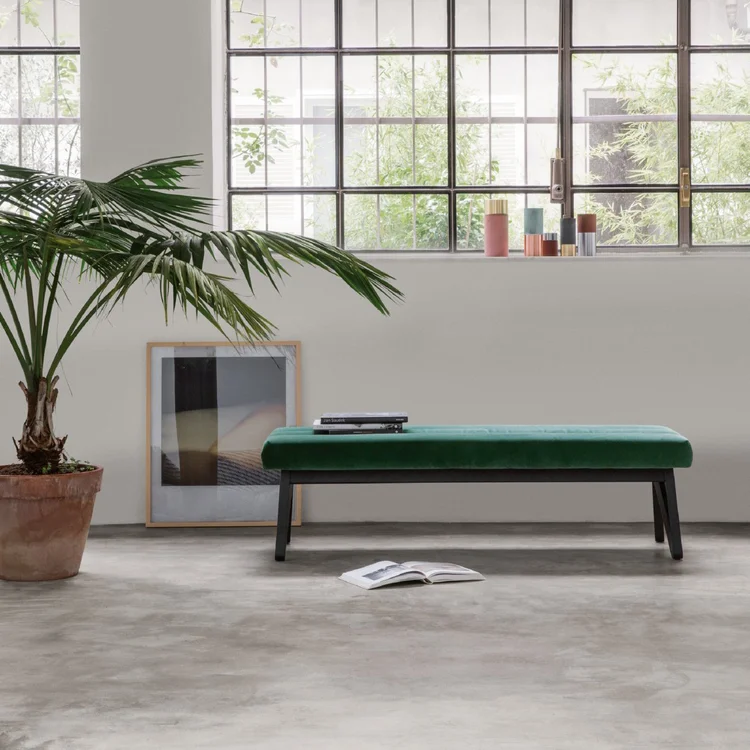 Modern interior with a green upholstered bench on black legs positioned against a white wall below large grid-patterned windows, a potted palm plant to the left, a framed abstract art piece leaning on the floor, and an open book on a polished concrete floor.
