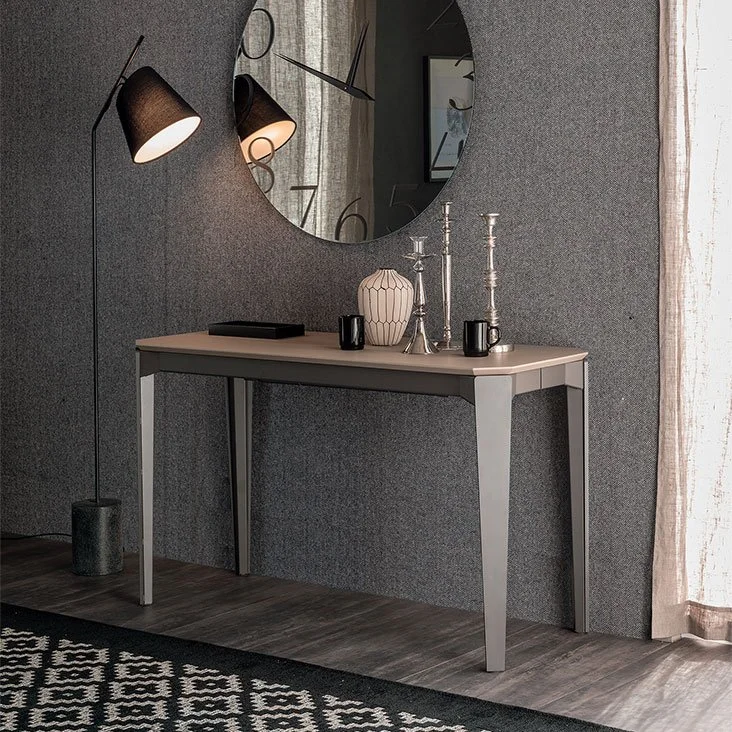 Modern minimalist console table with a light beige top and sleek legs against a textured grey wall, decorated with a ceramic vase, two black mugs, and three silver candlesticks, beneath a round wall clock with large numbers and accompanied by a tall floor lamp with two black lampshades and a patterned black and white rug on wooden floor.
