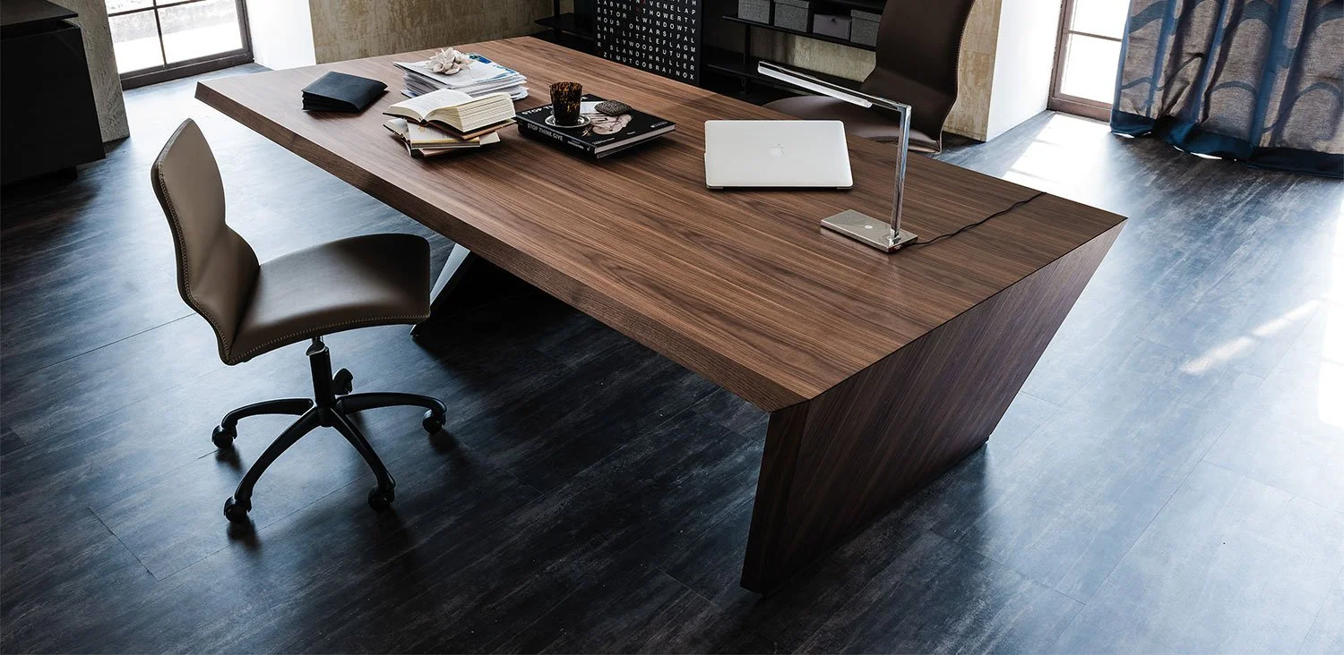 Modern office with a large wooden desk holding a laptop, books, notebooks, a lamp, and a glass cup, accompanied by a brown swivel chair on dark flooring.