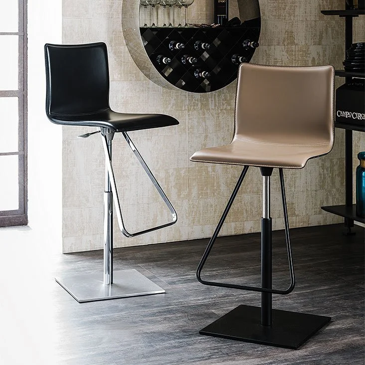 Two modern bar stools in a room with wood flooring and a light tiled wall, one stool with black seat and chrome legs, the other with tan seat and black legs, both with square bases and footrests.