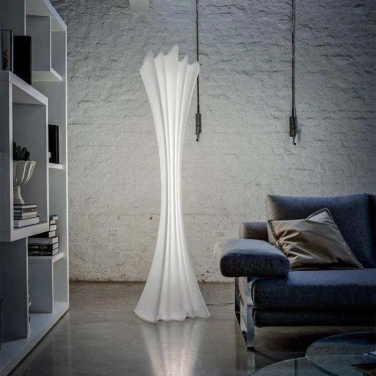 Modern white sculptural floor lamp with a flared, pleated design standing on a polished concrete floor in a living room next to a dark gray couch with pillows and a white bookshelf against a textured light gray wall.