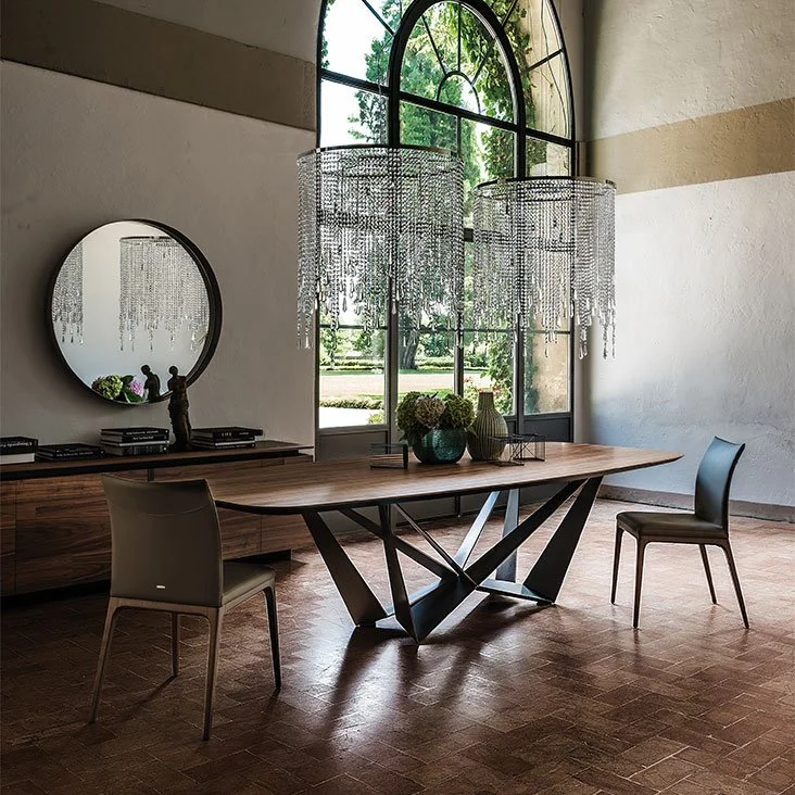 Modern dining room with a wooden table featuring a geometric black metal base and two gray chairs, under two large crystal chandeliers, beside a round mirror and a large arched window showing greenery outside.