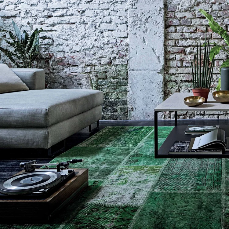 Modern living room with a light gray sectional sofa, green patterned area rug, coffee table with books and brass bowls, potted plants, and a vintage turntable on a wooden base against an exposed brick wall.