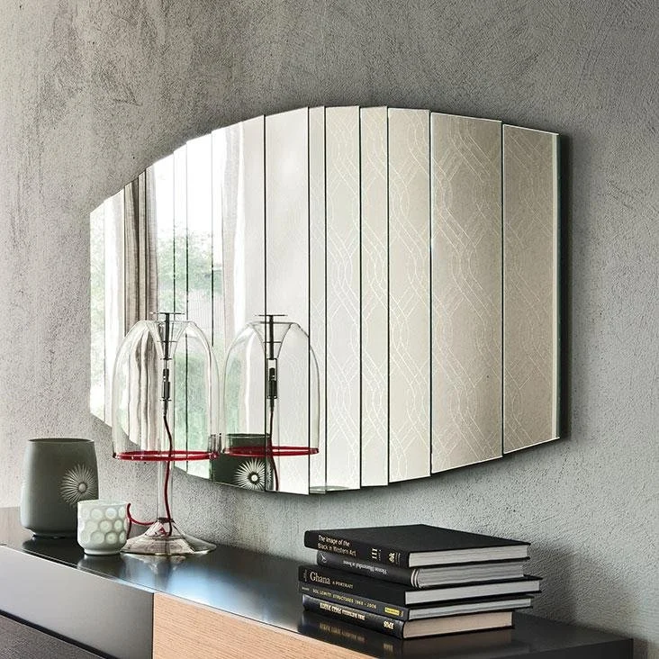 Decorative modern wall mirror with segmented panels mounted above a black and wood cabinet holding stacked books, a grey vase, and a glass lamp.