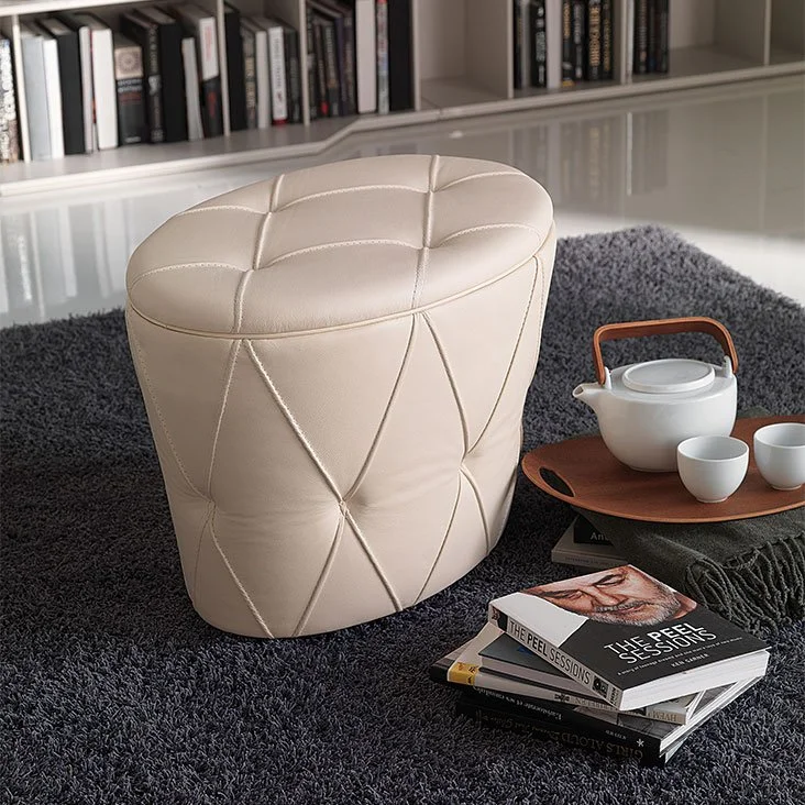 Cream-colored, oval-shaped tufted leather pouf on a dark gray shag rug next to a wooden tray holding a white teapot and two teacups, with a stack of books nearby and a bookshelf in the background.