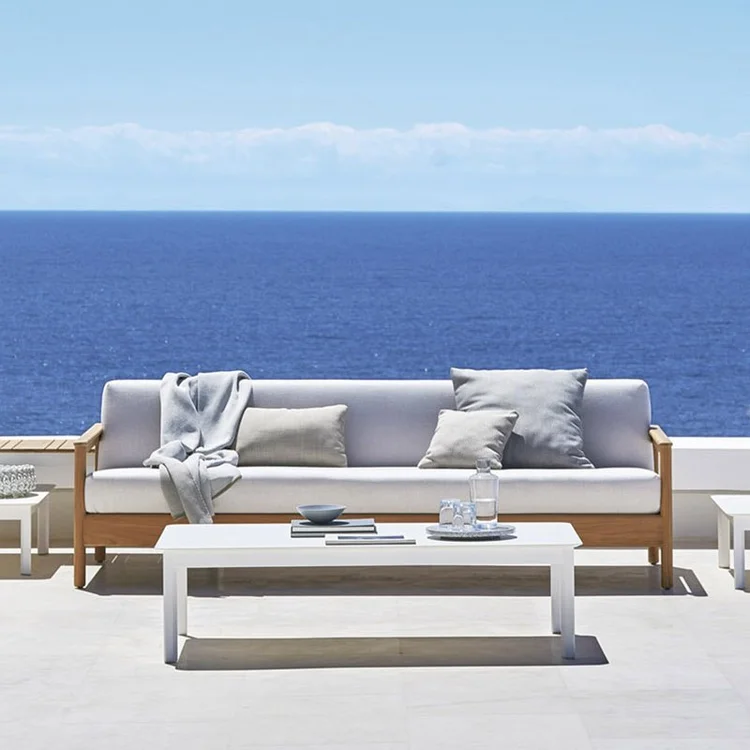 Outdoor seating area with a modern wooden sofa with light cushions and a throw blanket, white coffee table holding a carafe, glasses, and small dishes, overlooking a vast blue ocean under a clear sky.