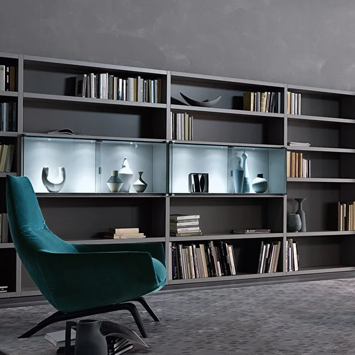 Modern living room with a large dark gray shelving unit holding books and ceramic vases displayed in glass-lit sections, and a teal upholstered armchair on a gray patterned carpet.