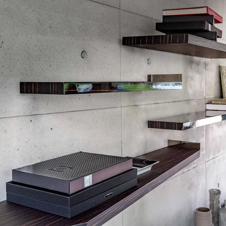 Minimalist wall-mounted dark wood shelves against a concrete wall, holding stacked books and decorative boxes.