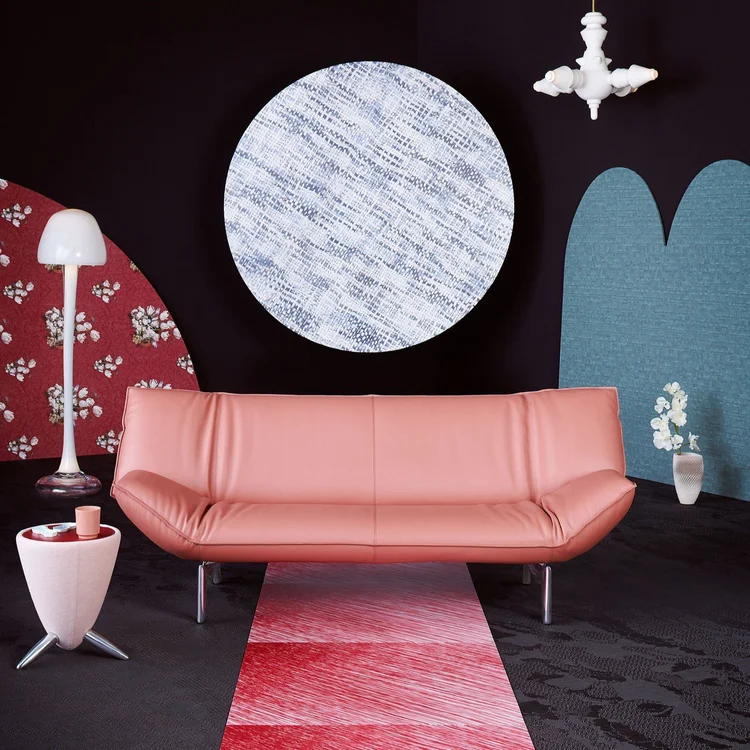 Modern interior with a pink leather sofa, a small triangular side table with a pink cup, a tall white floor lamp on the left, and a white vase with flowers on the right, all set against a dark wall with a large round patterned artwork and red and blue decorative panels.
