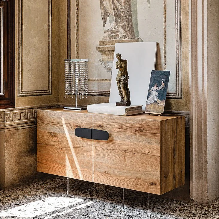 Wooden sideboard with black handle and clear legs placed against a wall with classical fresco artwork, decorated with a crystal table lamp, a small classical statue, a white book, and a small framed painting.