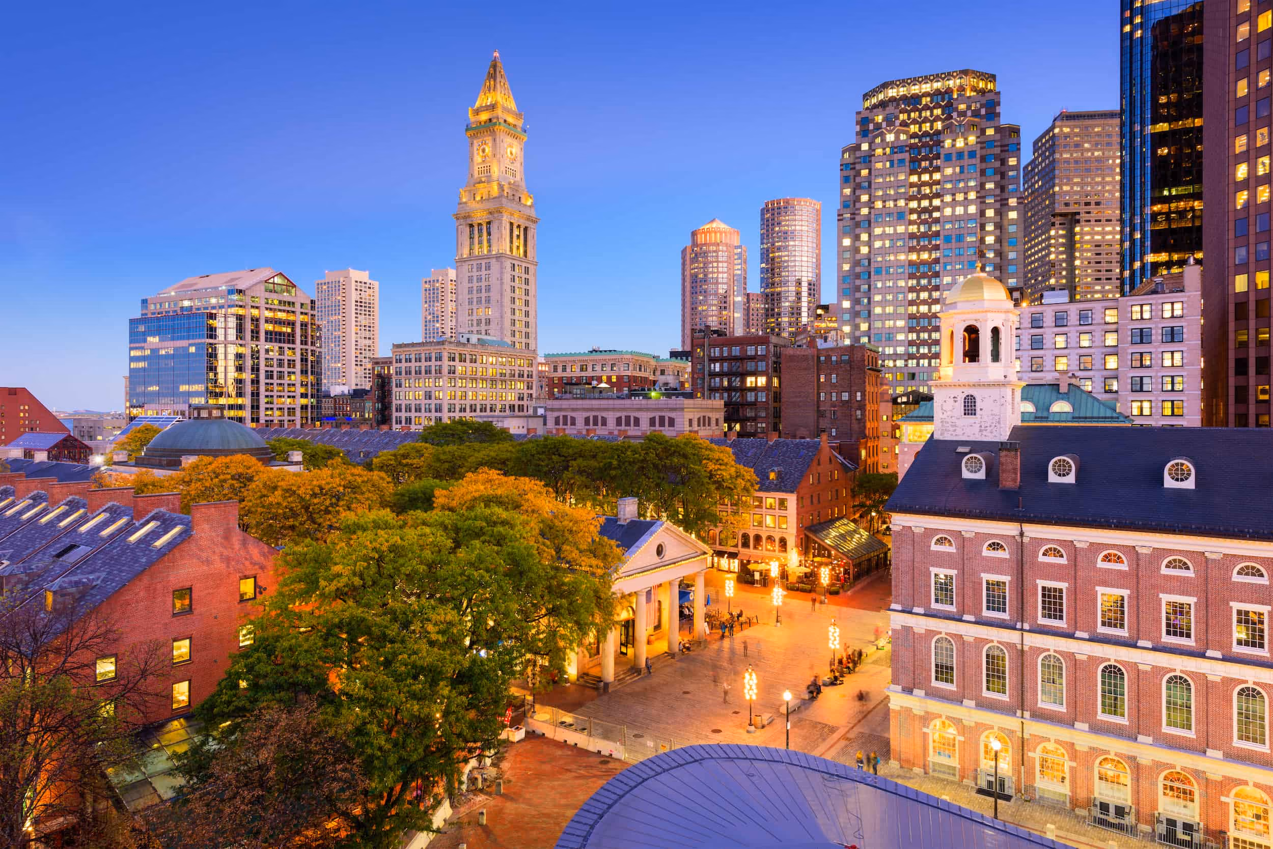 Evening cityscape of downtown Boston featuring illuminated historic and modern buildings, with trees and a plaza in the foreground under a blue sky.