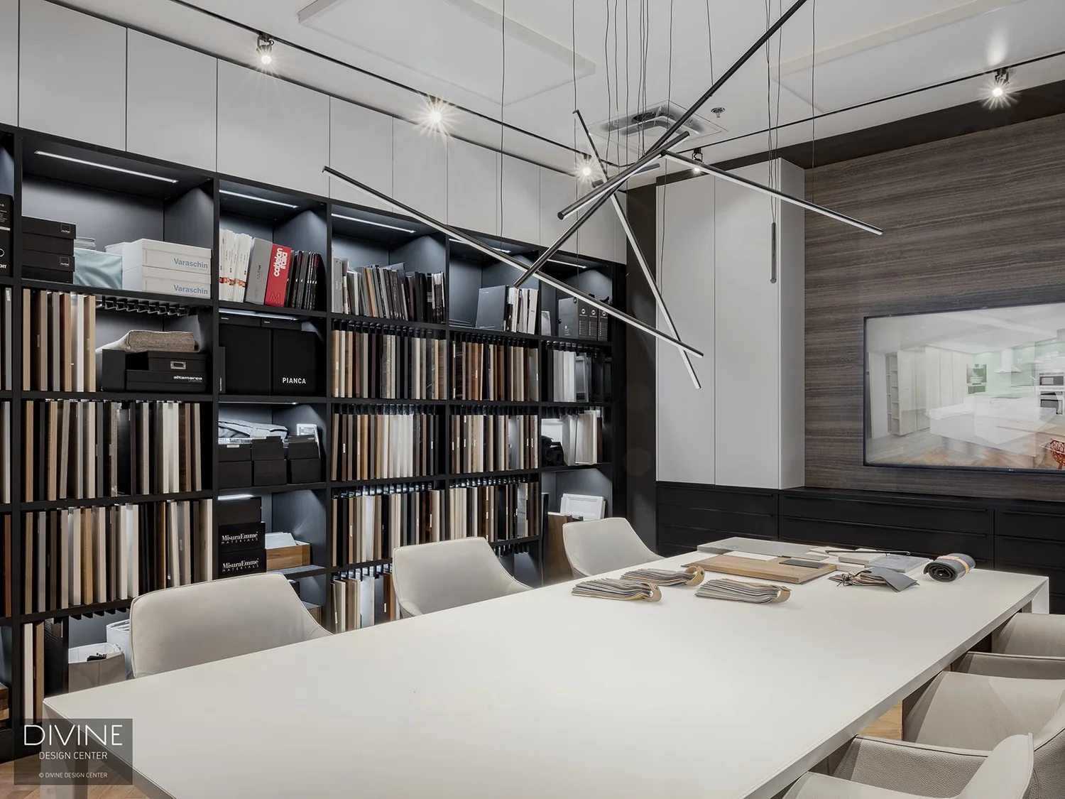 Modern design center meeting room with a large white table surrounded by beige chairs, a wall of shelves filled with samples and catalogs, and a mounted screen displaying an image of a kitchen. Suspended linear pendant lights hang above the table.