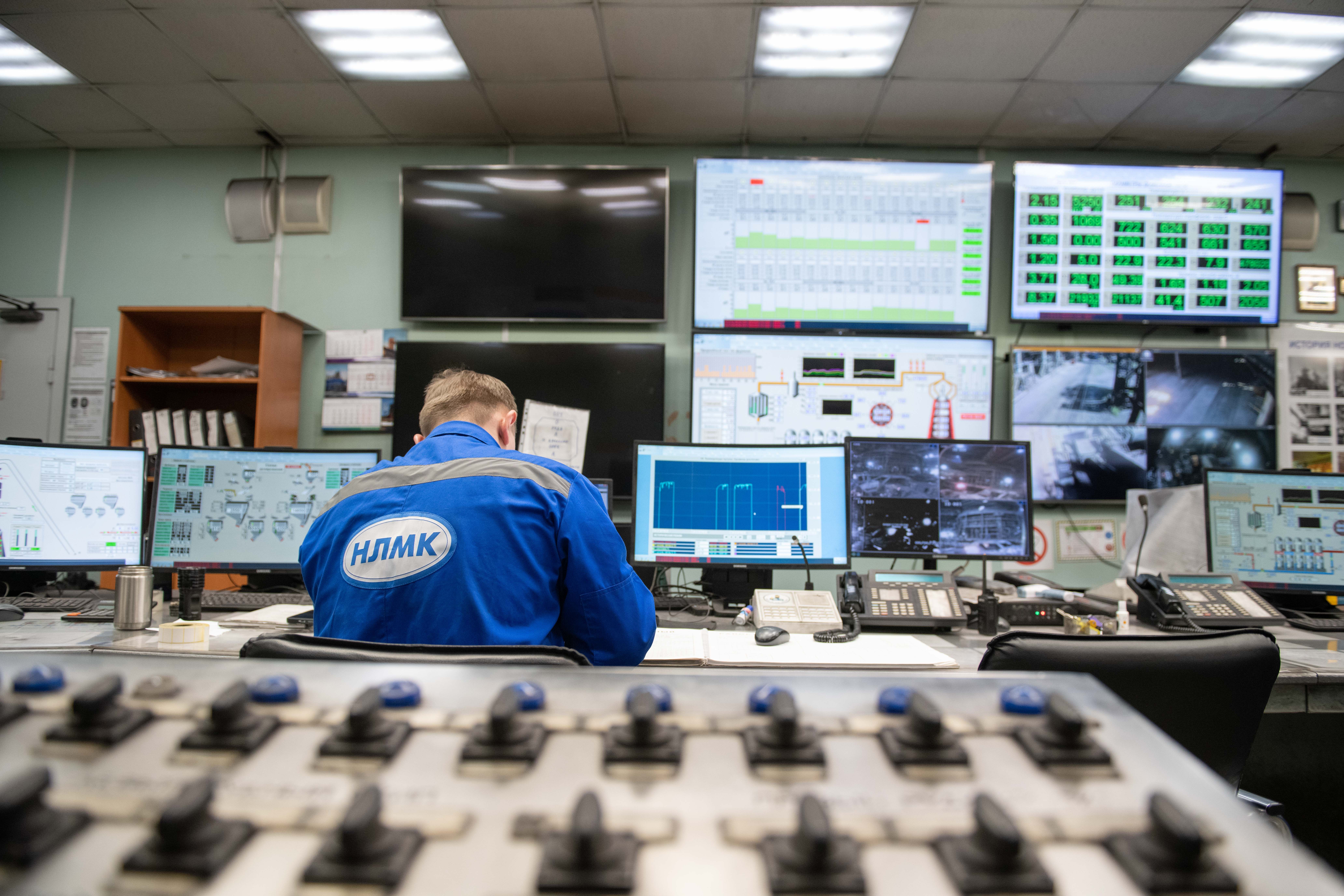 A control room with a wall-mounted screen split into multiple live camera feeds of the shop floor, and a person seated at a desk in front of it.
