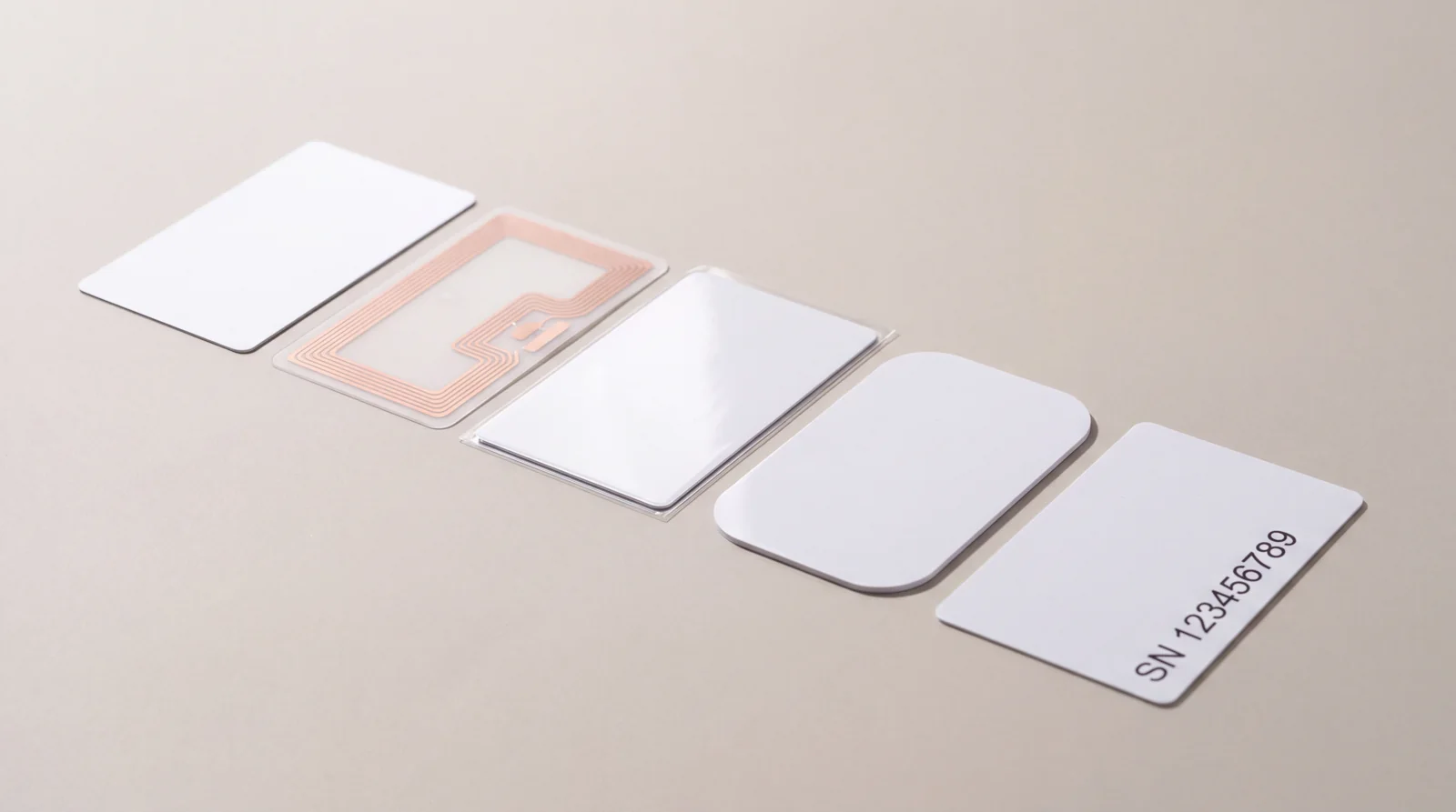 Five cards in sequence showing plastic card production stages, from a blank card and a copper RFID antenna inlay to a laminated card, a die-cut shape and a numbered finished card