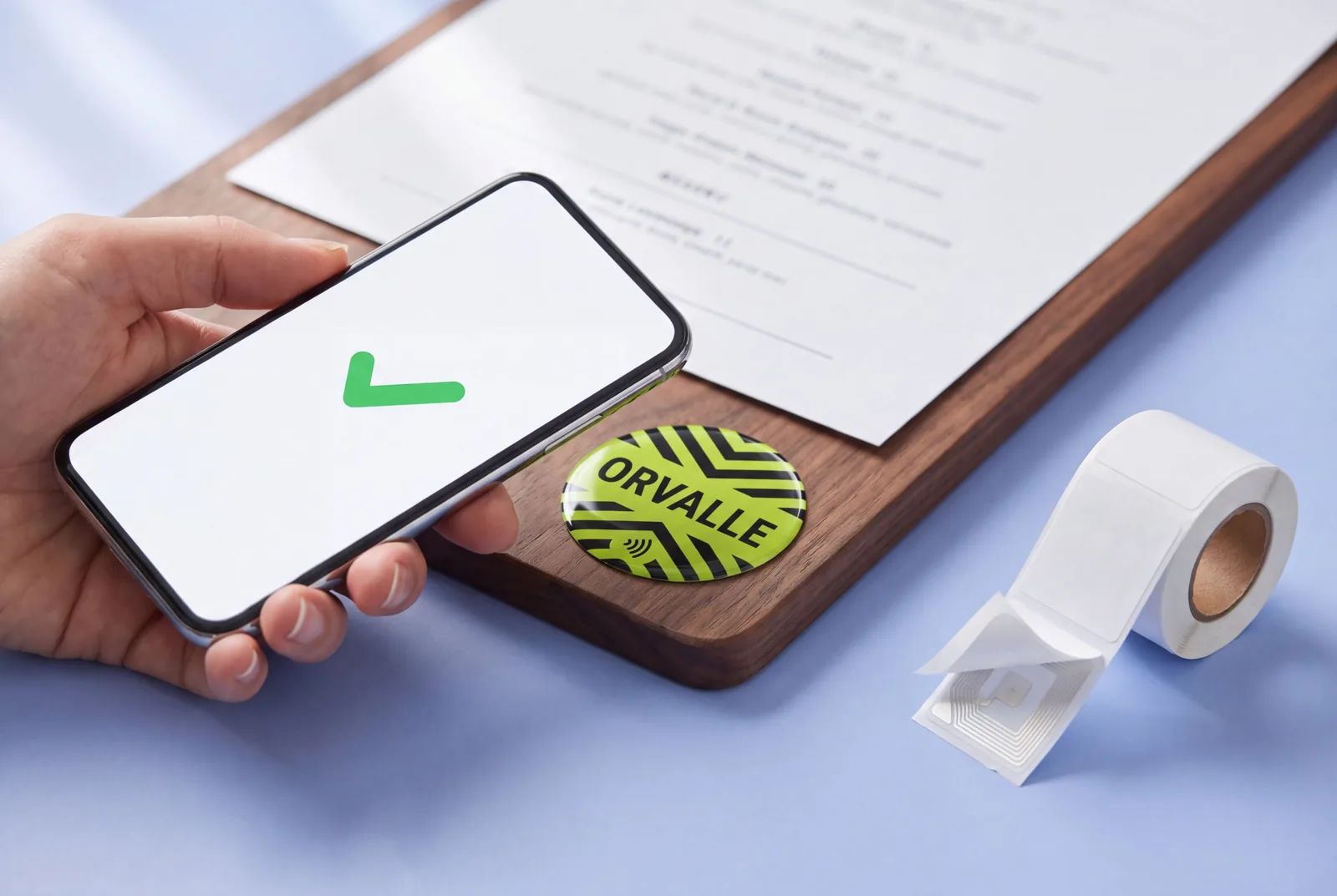 A hand holding a smartphone over a round lime and black NFC sticker for a fictional restaurant on a wooden menu board, beside a roll of RFID labels with the antenna visible