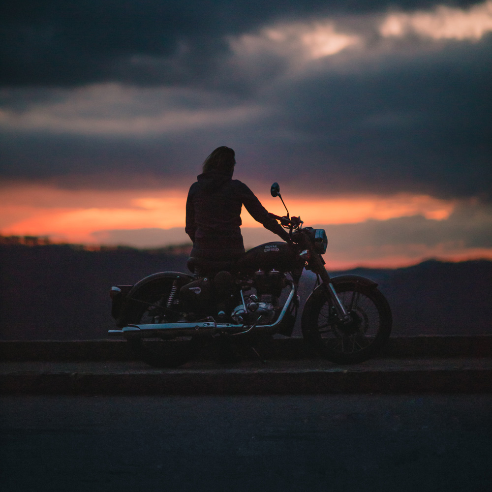 Silhouette of a person sitting on a Royal Enfield motorcycle during sunset with a dark cloudy sky in the background.