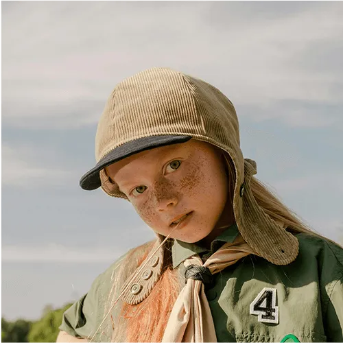 Young girl with freckles wearing a beige corduroy cap and green outfit with a neckerchief and number 4 patch, standing outdoors against a cloudy sky.