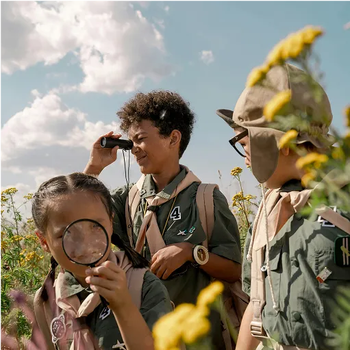Three children in scouting uniforms outdoors in a field with yellow flowers; one girl in front holds a magnifying glass up to the camera, a boy in the middle looks through binoculars, and another child stands on the right wearing a hat.