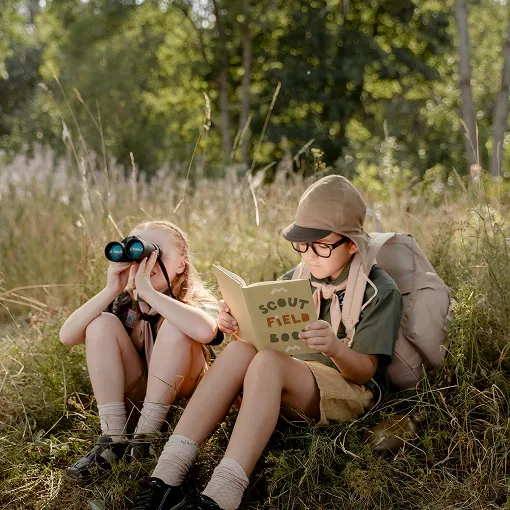Two children sitting on grass in a forested area; one child looks through binoculars while the other reads a book titled 'Scout Field Book'. Both wear outdoor gear including hats and backpacks.
