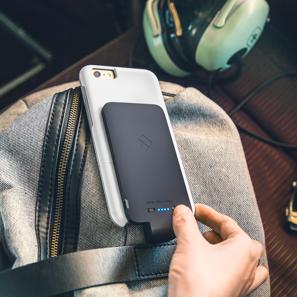 Hand holding a white smartphone with a black Polar Pro accessory attached on its back, resting on a grey backpack with a black leather strap and zipper, with green headphones and a car key in the background.