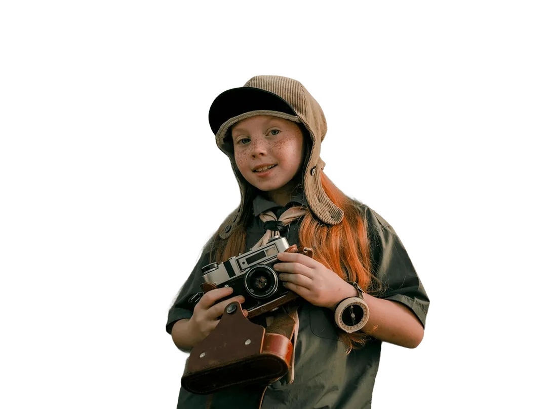 Young girl with long red hair, freckles, and a green scout uniform wearing a brown aviator-style hat and holding a vintage camera in a brown case.