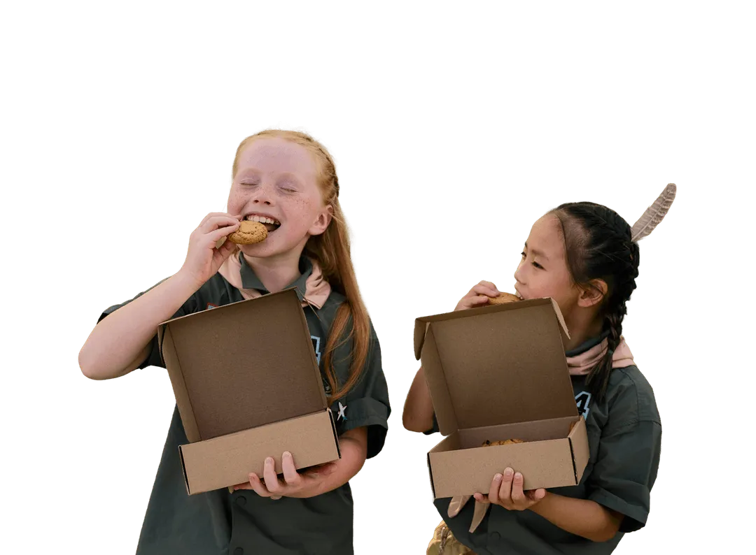 Two girls in scout uniforms eating cookies from open cardboard boxes, one girl with red hair smiling and eyes closed, and the other girl with braided hair and a feather in her hair, looking at the cookies.