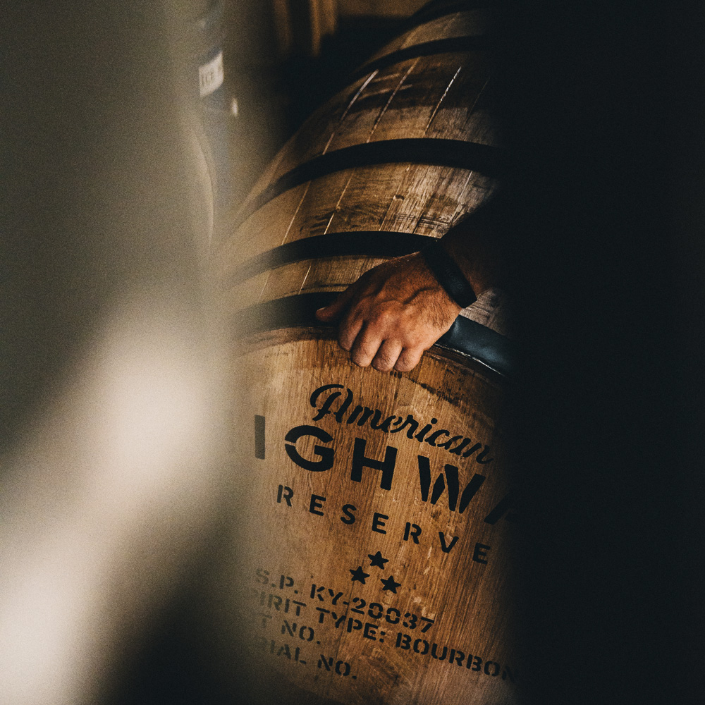 A person's hand with a wristband resting on a wooden barrel labeled American Highway Reserve with bourbon spirit type and batch details, partially obscured by dark shadows.