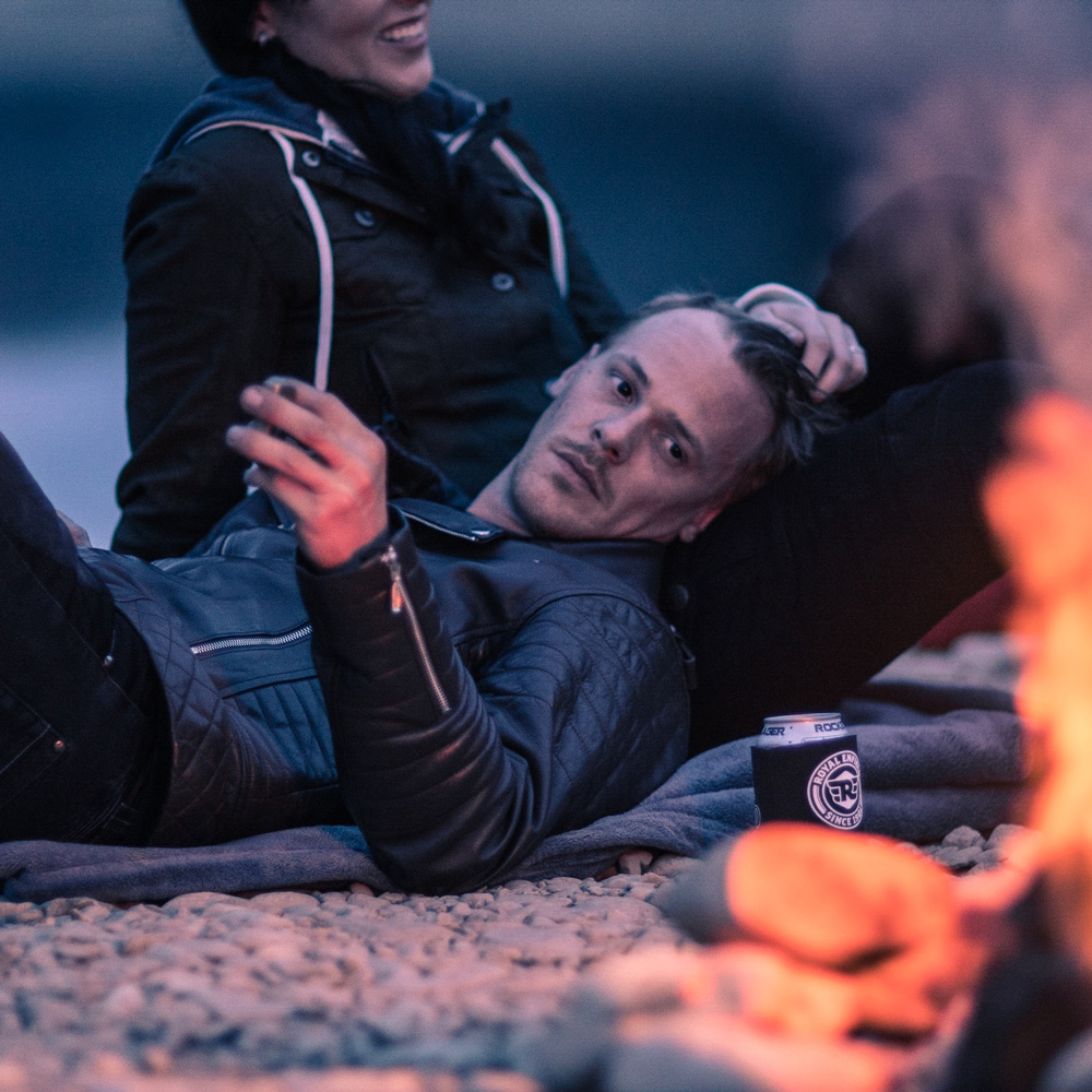 A man in a black leather jacket lies on a blanket on a pebble beach, resting his head on a woman's lap; a fire burns nearby, and a can of Royal Empire beer is visible beside them.