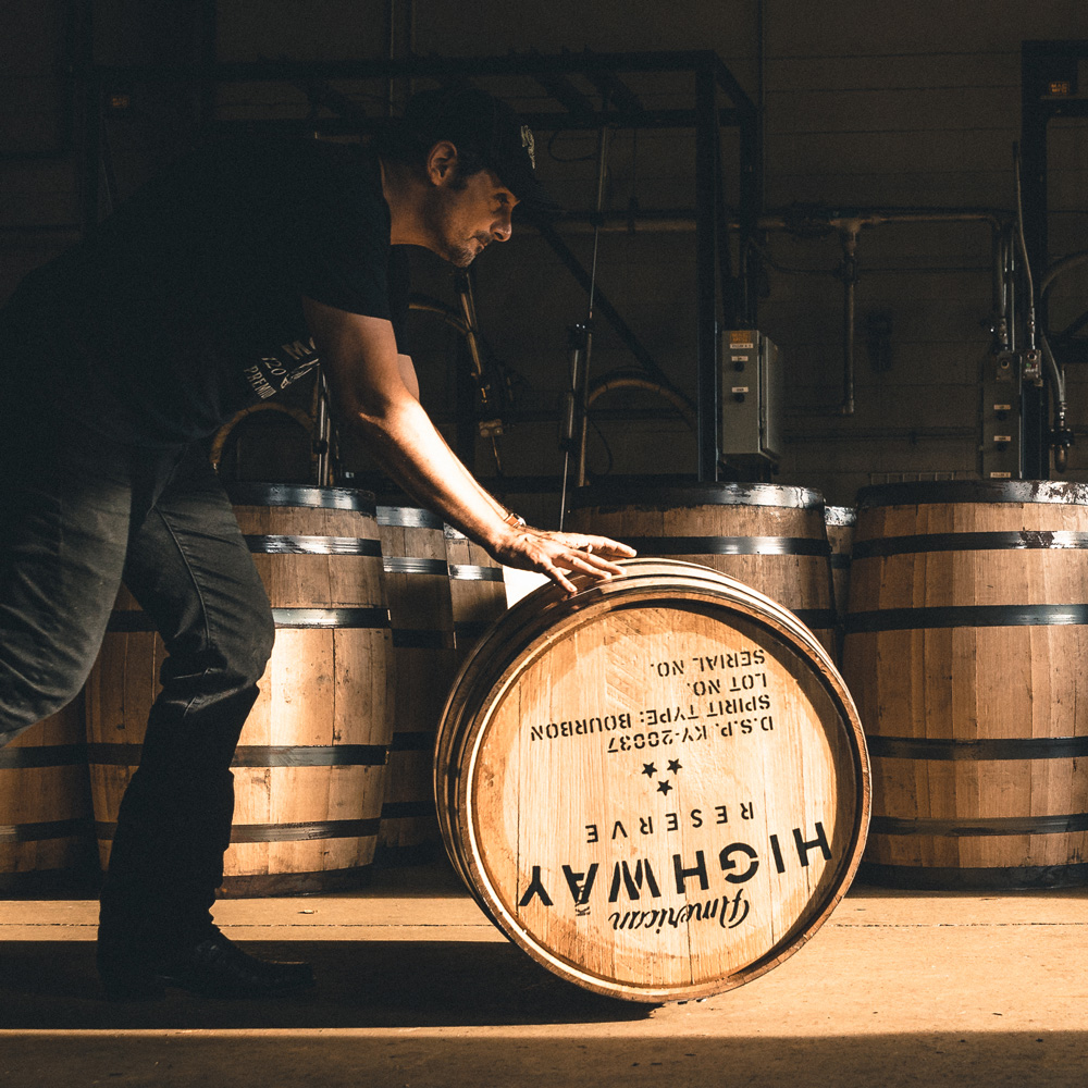Man wearing dark clothes and cap rolling a wooden barrel labeled 'American Highway Reserve' inside a dimly lit warehouse with other barrels in the background.