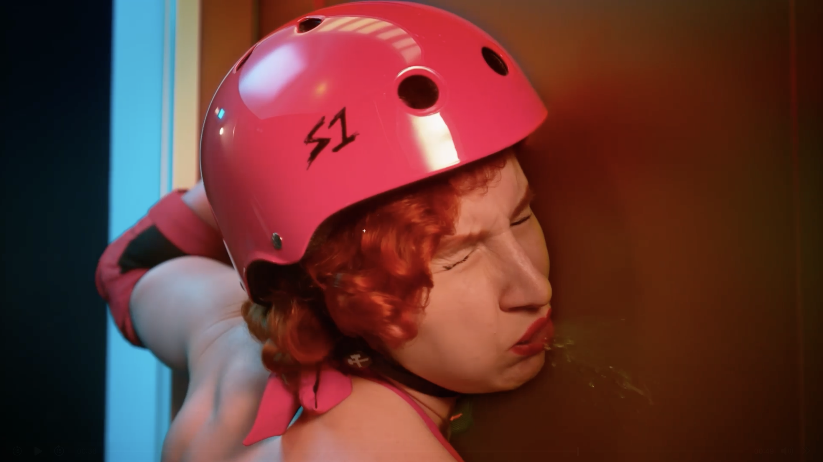 Person with curly red hair wearing a bright pink helmet with 'S1' on it, grimacing and appearing to spit against a reddish-brown wall.