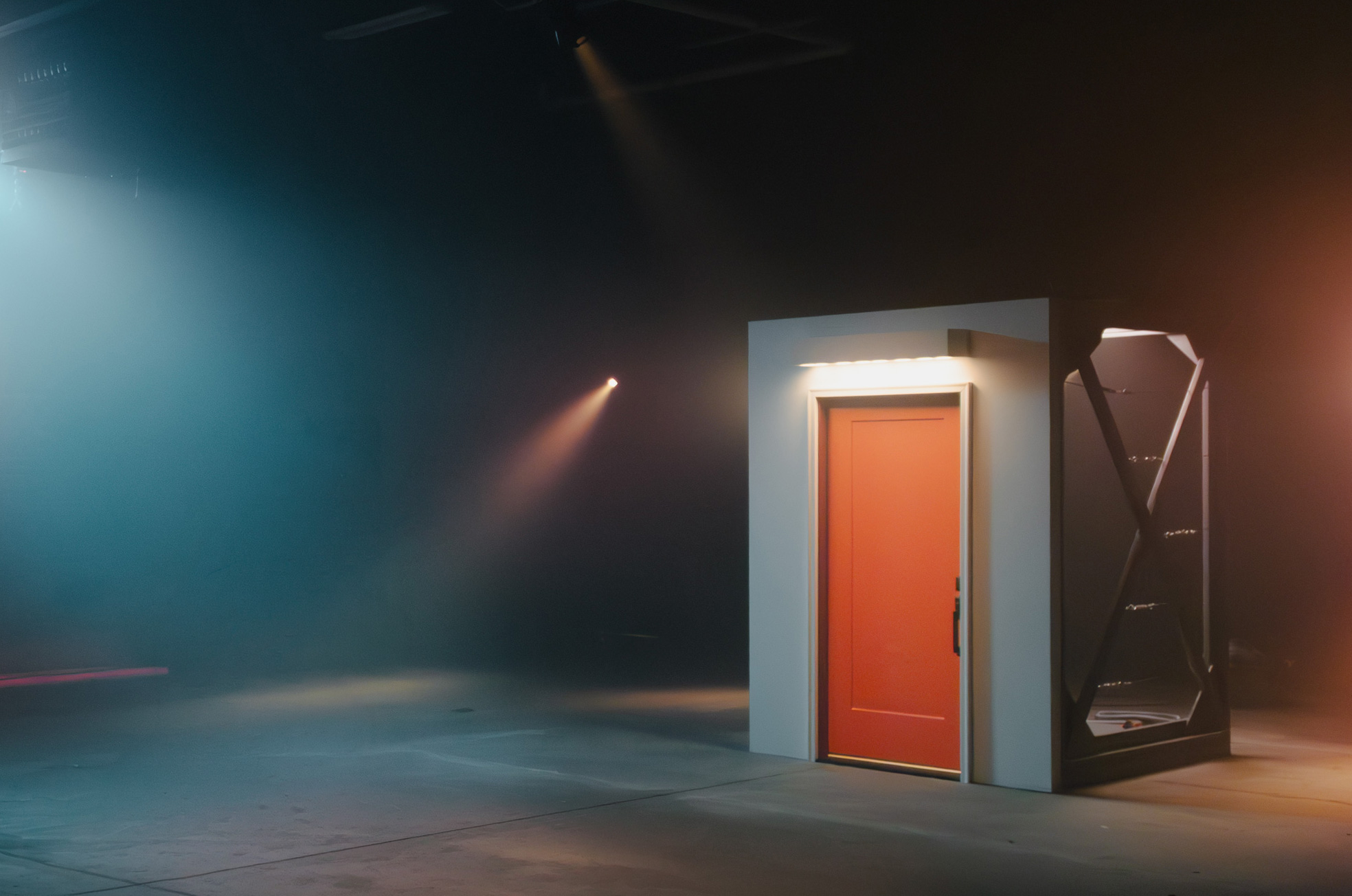 Isolated small white structure with a closed red door in dimly lit, foggy space with spotlights and concrete floor.