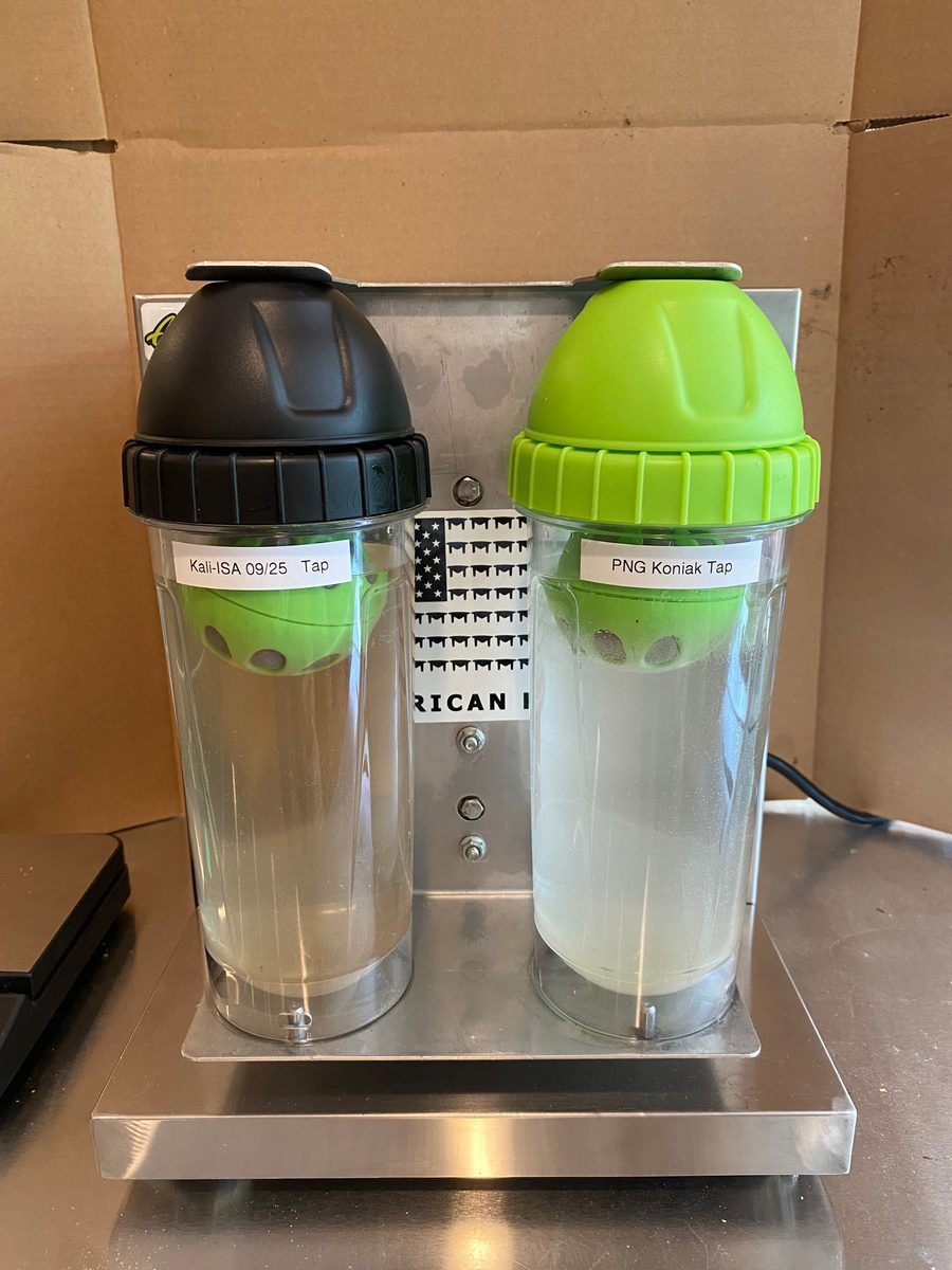 Two labelled Alu Shaker Pro bottles of clear water loaded into the brackets of the Alu Bottle Shaker machine before a run
