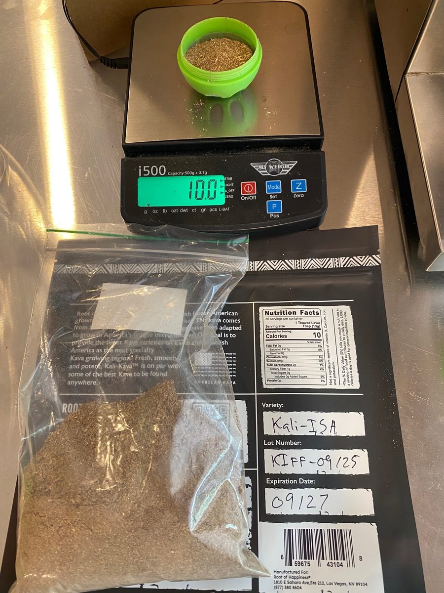 A green Alu Ball straining vessel on a digital scale reading 10.0 grams of milled Kali-ISA kava powder, with a labelled sample bag alongside