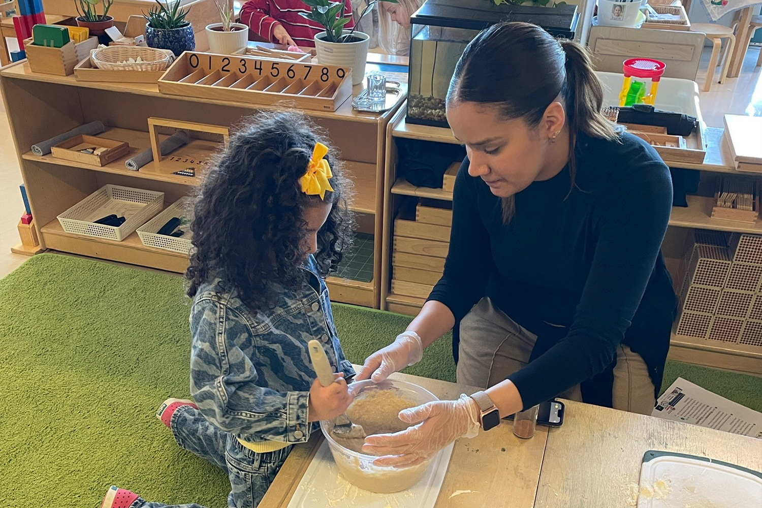Battery Park Montessori student mixing materials with a paintbrush while the teacher holds the bowl.
