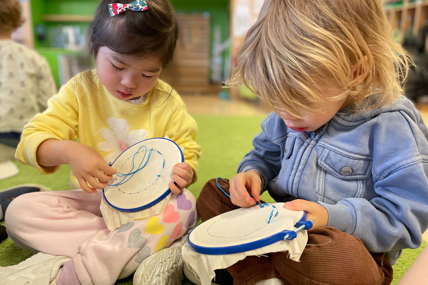 As part of Montessori's Practical Life focus, two students practice their sewing skills on an embroidery hoop.