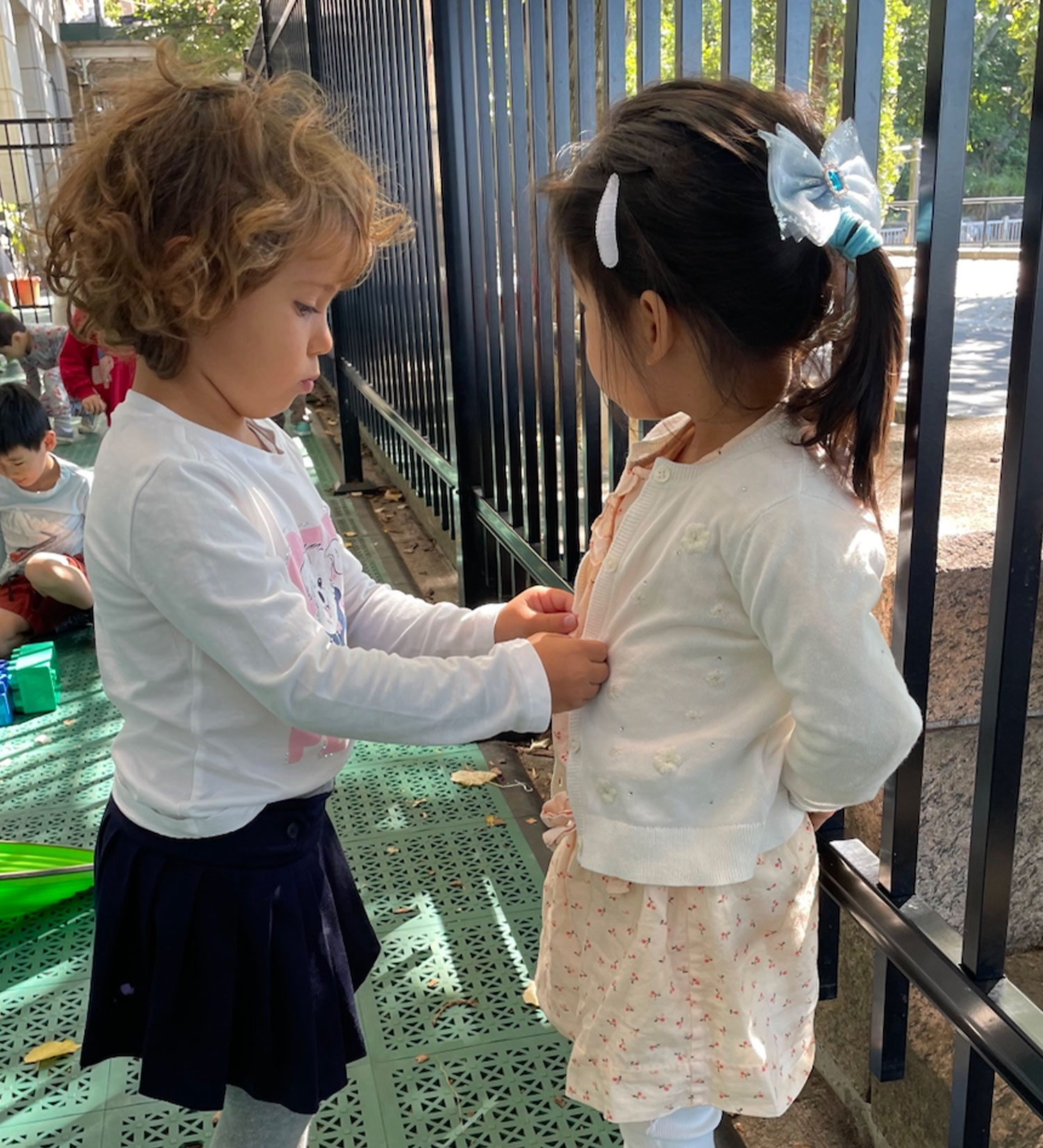 An older student helping a classmate button her cardigan as an example of leadership and mentorship in a mixed age Casa classroom