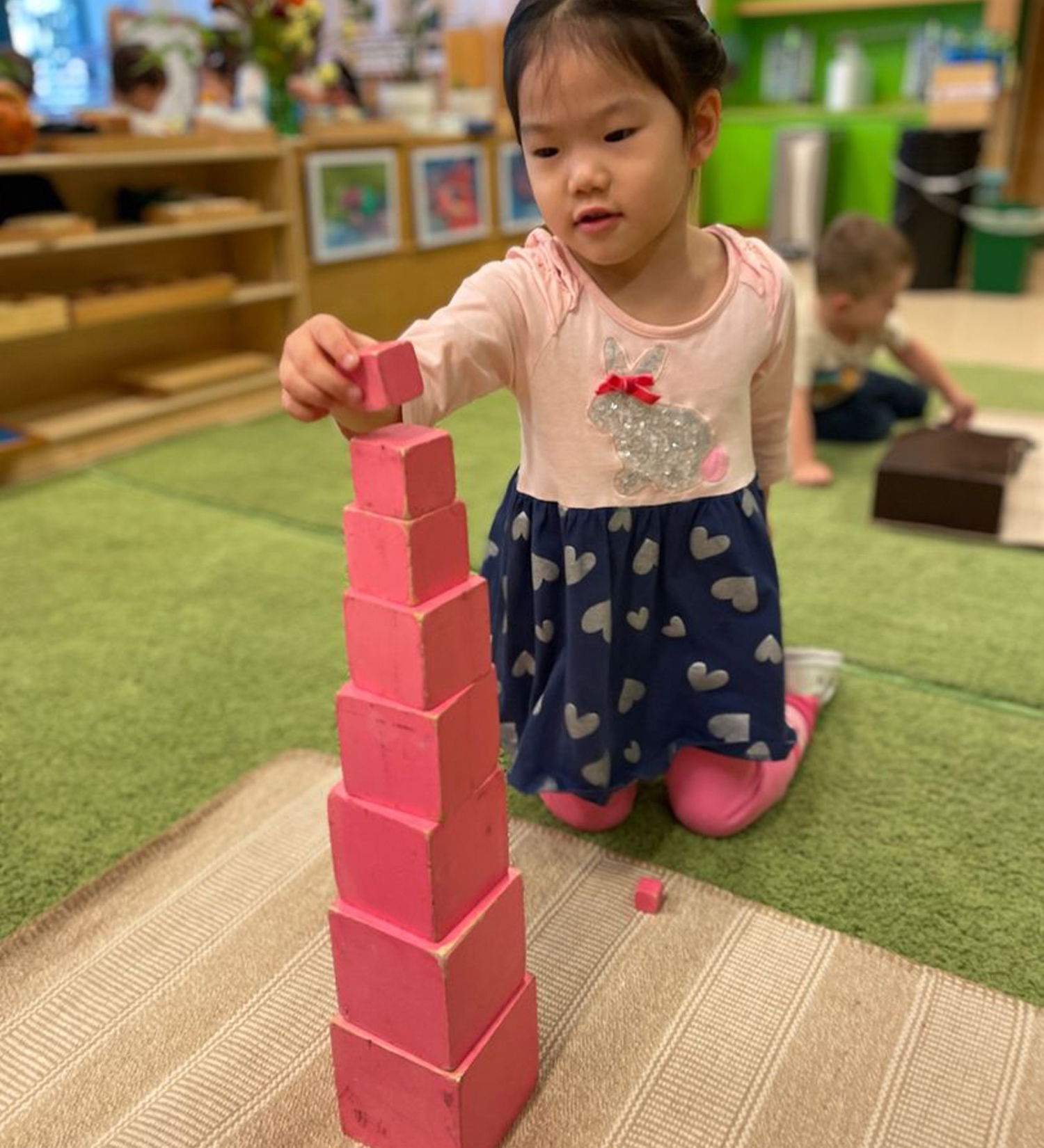 A student stacking the Montessori Pink Tower, a classic sensorial material featuring 10 graduated pink wooden cubes that children stack from largest to smallest.
