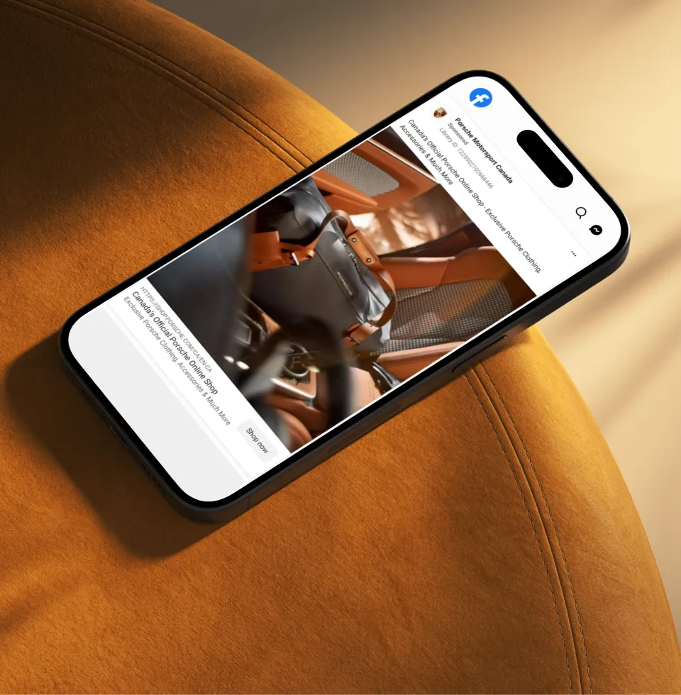 Smartphone lying on an orange suede surface, displaying a Facebook page of Porsche Motorsport Canada featuring a stylish black and brown leather Porsche bag with orange details inside a car.