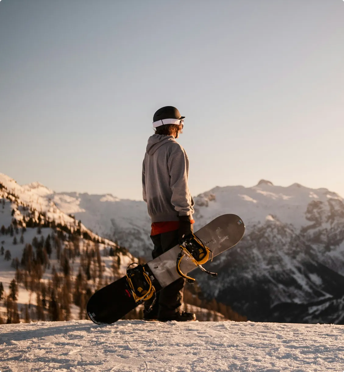 Person dressed in winter clothing and helmet holding a snowboard, standing on snow with a backdrop of snow-covered mountains at sunset.