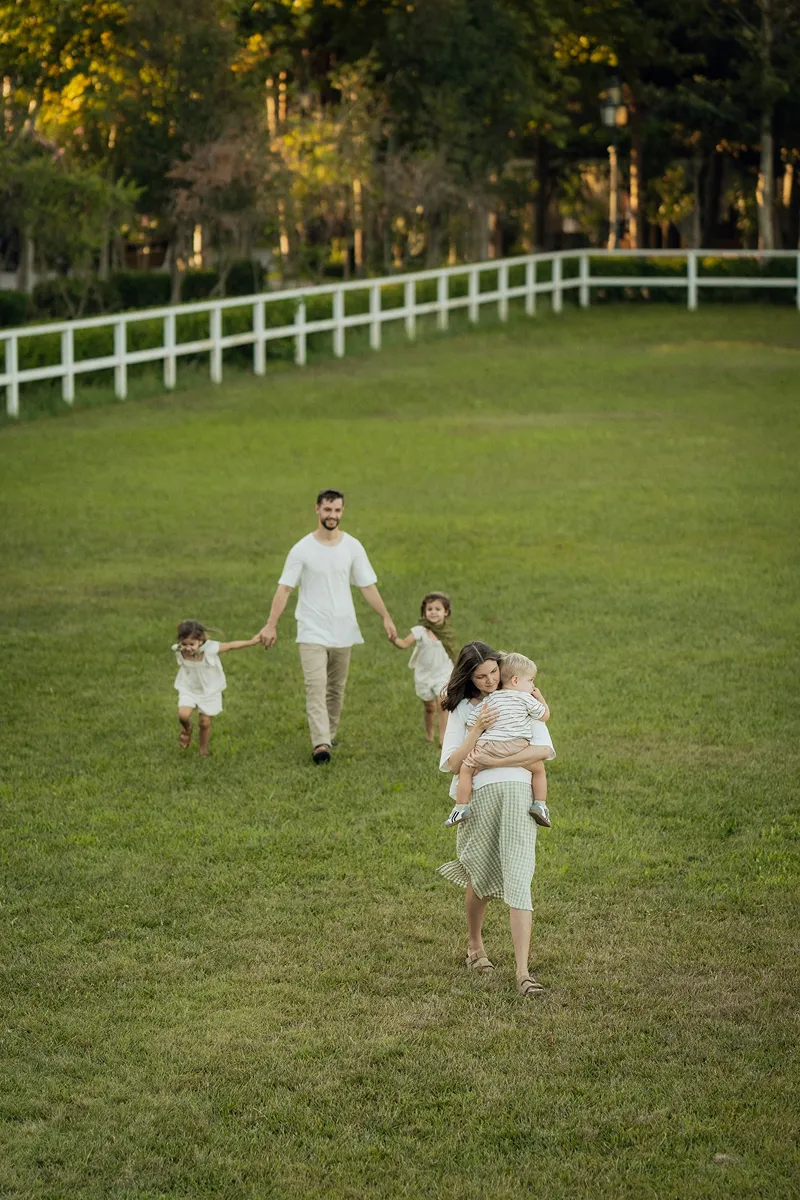 A family of five outdoors on a grassy hill near a white fence; a woman walks in front carrying a toddler, while a man behind holds hands with two girls.